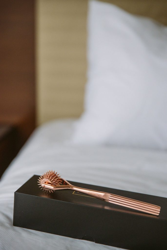 The rose gold pinwheel sits out on top of its black box on top of a bed with white bedding. The headboard is visible in the background, and the spikes of the pinwheel look extremely sharp. For my Liebe Seele X-Rose Wartenburg Pinwheel review.