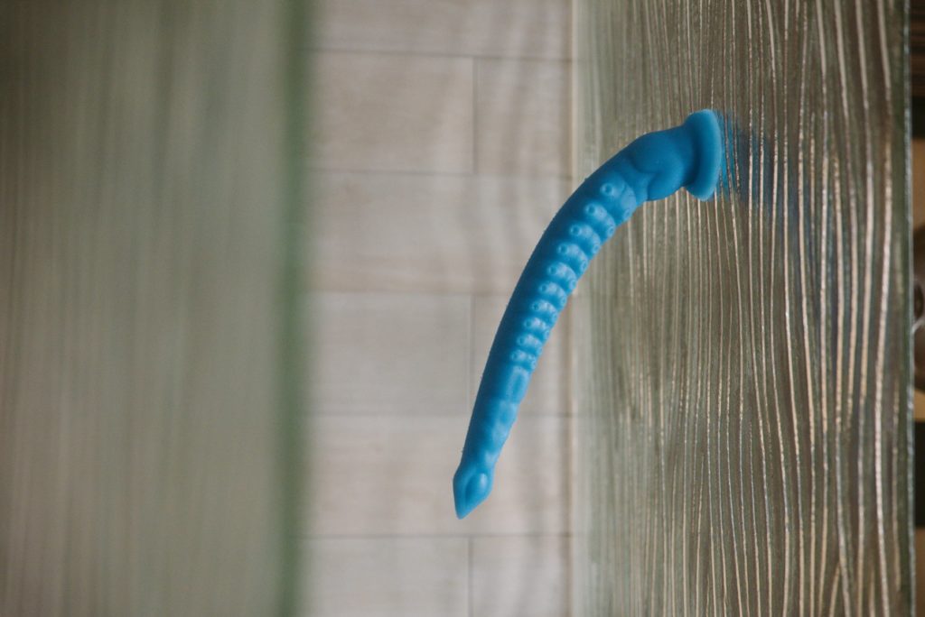 A blue tentacle dildo is stuck to a glass, textured shower wall. It drapes with the pull of gravity, but it doesn't collapse entirely sideways. It looks very textured with a lot of little tentacle suckers. For my MEO.de Long Anal Tentacle Dildo review. 