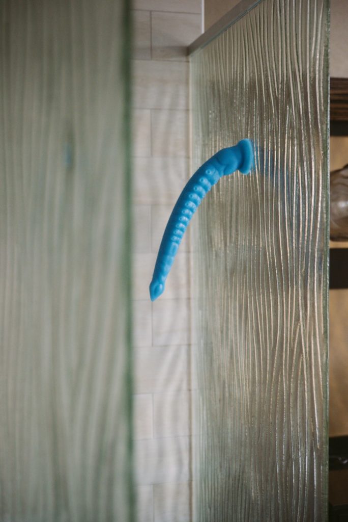 A blue tentacle dildo is stuck to a glass, textured shower wall. It drapes with the pull of gravity, but it doesn't collapse entirely sideways. It looks very textured with a lot of little tentacle suckers. For my MEO.de Long Anal Tentacle Dildo review.