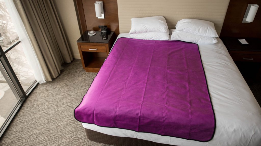 The Aubergine Purple Liberator Throw lays out on a King-sized hotel room bed. It covers about 3/4th of the width of the bed and almost the entirety of the length of the bed. It looks vibrant and welcoming. For my Liberator Throw review.