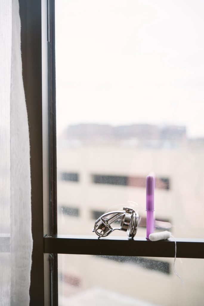 A chastity cage and a purple tampon sit out on a windowsill, looking well-lit as the focus of the entire frame. Both look welcoming in the natural light. For my Menstrual Period Chastity 101 article.
