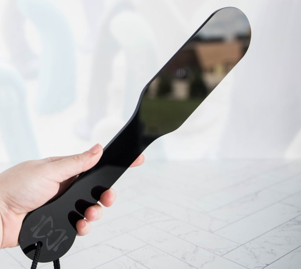 My hand wrapped around the handle of the Sincerely Sportsheets paddle. The mirrored surface showcases other colors from the room. For my Sportsheets Sincerely Bundle review.
