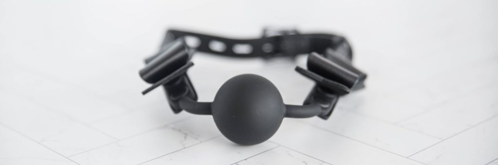 Front view of the Sportsheets Sincerely ball gag laying on a tiled surface. It showcases the round, silicone, plushy-looking ball of the gag itself as well as the two silicone, slim "arms" that extend outwards from the ball to fasten to the faux leather head strap. For my Sportsheets Sincerely Bundle review.