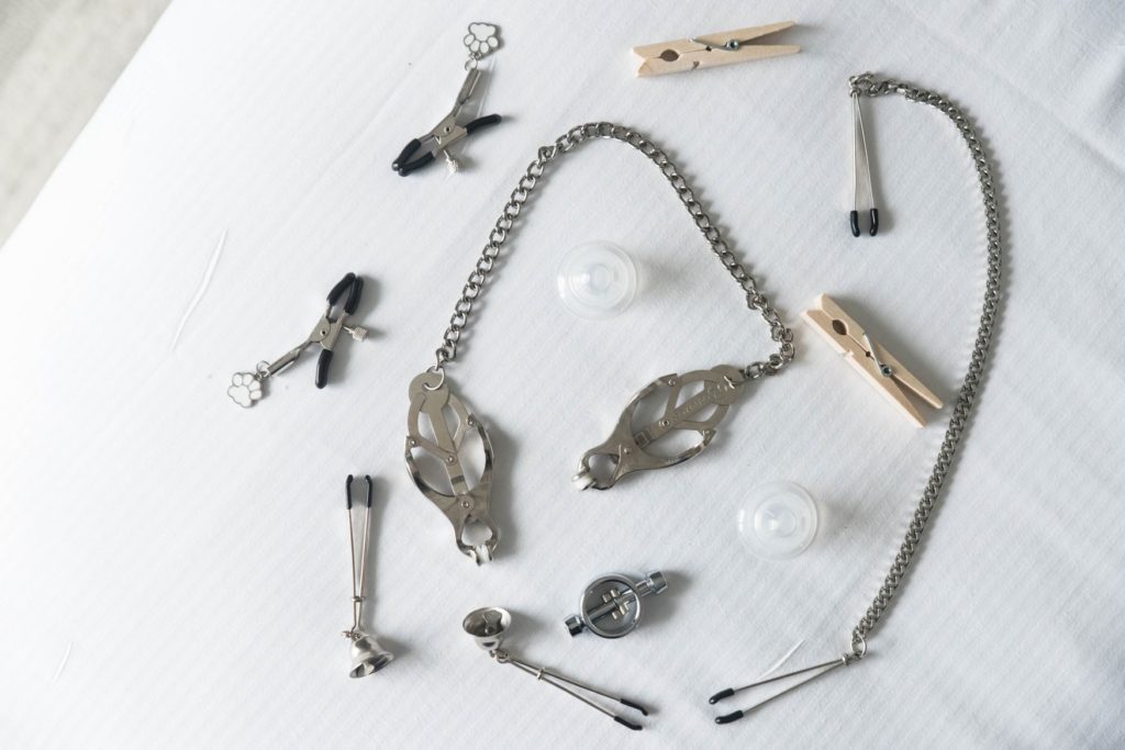 A whole collection of nipple clamps laid out on a white bed spread, showing a whole host of different types of nipple clamps. For my BDSM Nipple Clamps article.