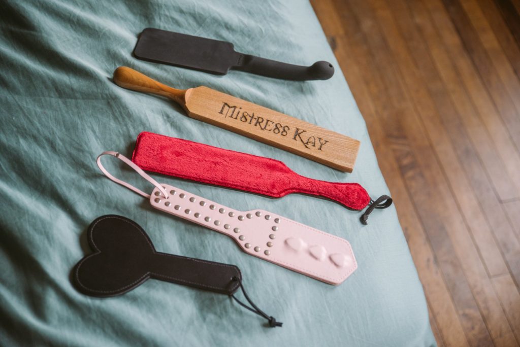 A spread of kinky paddles laying out the blue bedspread, showcasing their different sizes and colors and styles. For my Everything You need to Know about Using a BDSM Paddle review.