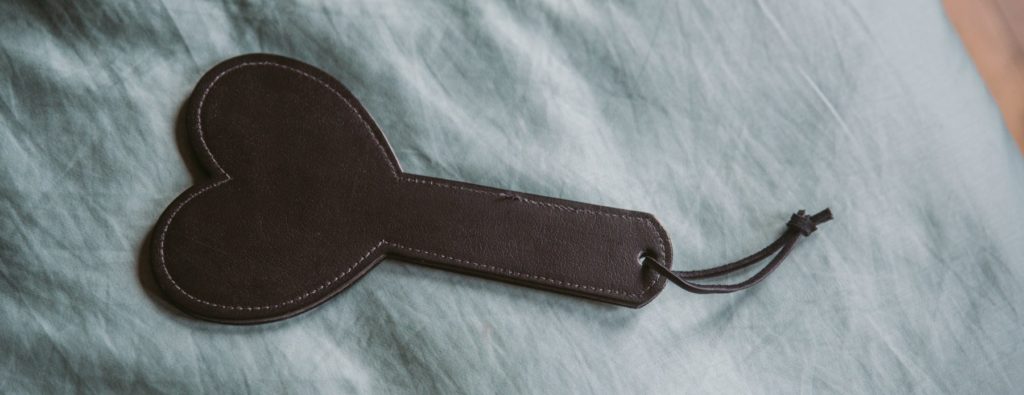 A black, heart-shaped faux leather paddle laying on top of a blue bedspread.