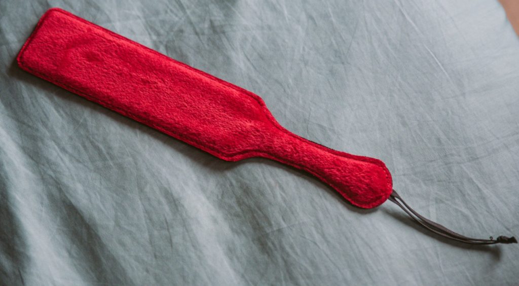 A double-sided paddle with the red faux fur side showing, laying on top of a blue bedspread.