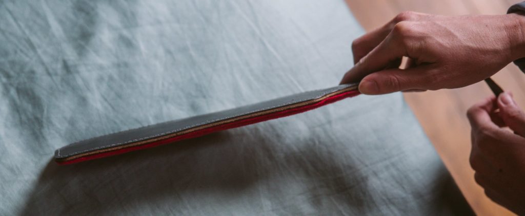 A person holds a double-sided paddle to the camera with the edge of the paddle facing this camera. This angle showcases the thin, black leather layer on one side and the red faux fur layer on the opposing side. For my Everything You need to Know about Using a BDSM Paddle review.