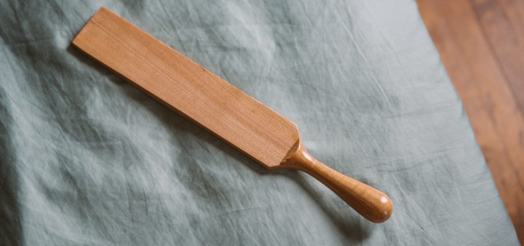 A wooden paddle laying on top of a blue bedspread. For my Everything You need to Know about Using a BDSM Paddle review.
