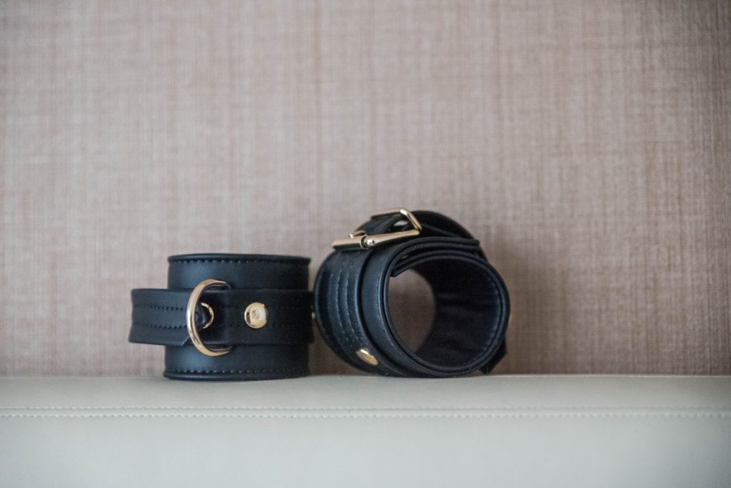 The two black cuffs lay out on top of white upholstery. A cross-stitch wall is in the background. The cuffs look regal and fancy with their contrasting gold hardware. For my Liebe Seele Dark Candy Cuffs review.
