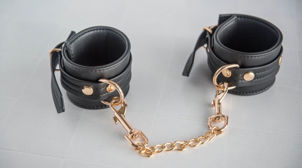 Both of the cuffs sitting out on a tiled surface. The double-sided clip chain that's included is shown clipped onto both of the cuffs, turning them instantly into handcuffs that can be used right away for bondage. For my Liebe Seele Dark Candy Cuffs review.