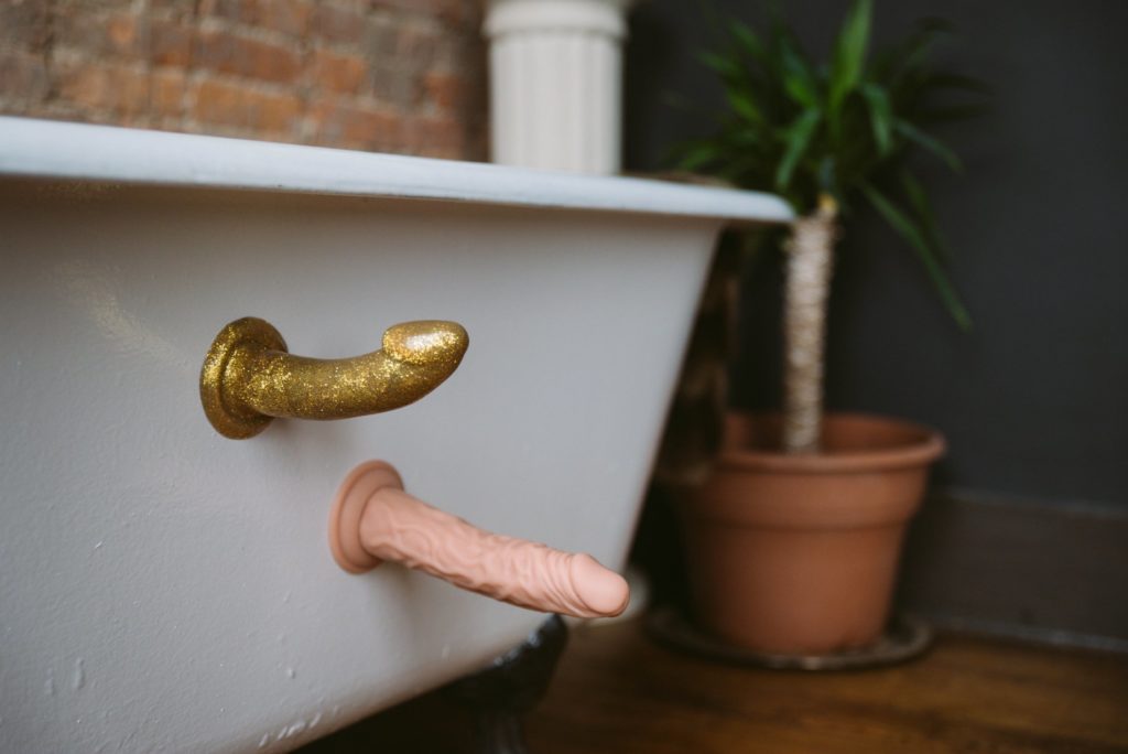 Two suction cup dildos are stuck onto the side of my bathtub. One is realistic and one is sparkly and gold. Both are staying on the side of the bathtub with nothing else to keep them there. Feature image for my How to Use a Suction Cup Dildo article.