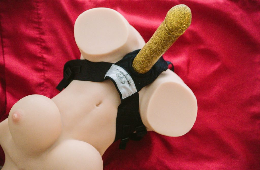 A sex doll is laying on top of a red, satin background. A strap-on harness is wrapped around the sex doll's waist with a gigantic golden dildo protruding from the doll on the harness. Image for my How to Cuckold for Beginners: Using a Sex Doll.