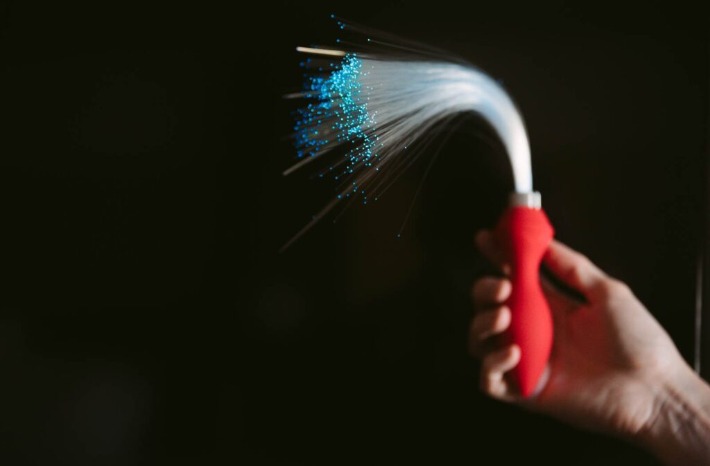 A hand holds fiber optic anal plug against a black background. The fiber optics are turned on, and the end of the tail glows brightly with the color of the fiber optics. For my Inyarose Luminous Fiber Optic anal plug review.