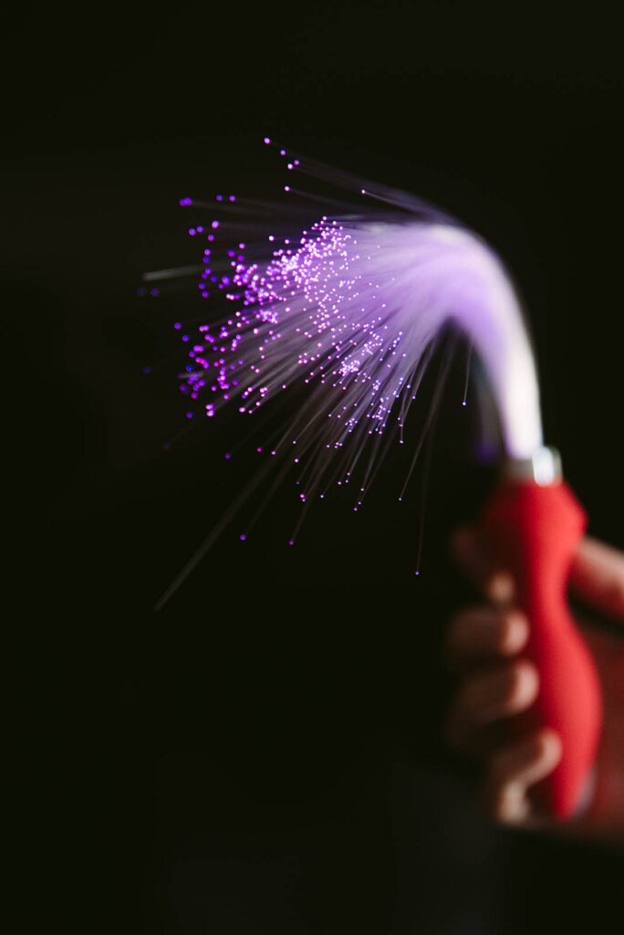 A hand holds fiber optic anal plug against a black background. The fiber optics are turned on, and the end of the tail glows brightly with the color of the fiber optics. For my Inyarose Luminous Fiber Optic anal plug review.