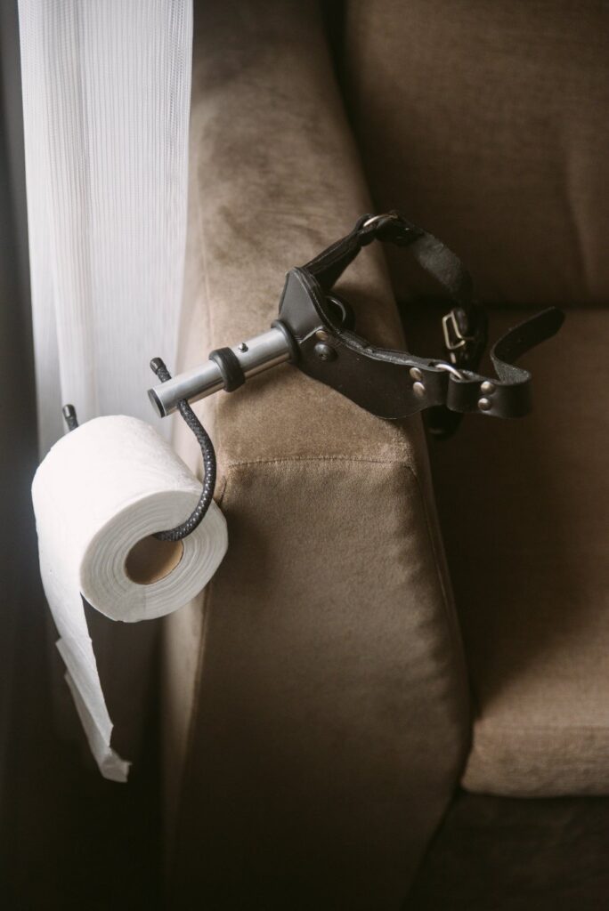 The gag with attached toilet paper attachment hangs off a chair, holding a toilet paper roll. Image for my Getting Ready for a Femdom Date BDSM Scene