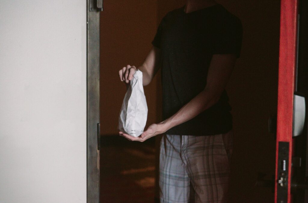 A person is standing in an open, shadowy door, holding a bag of food. It looks like a delivery driver delivering food. Image for my easy home sex roleplay ideas.