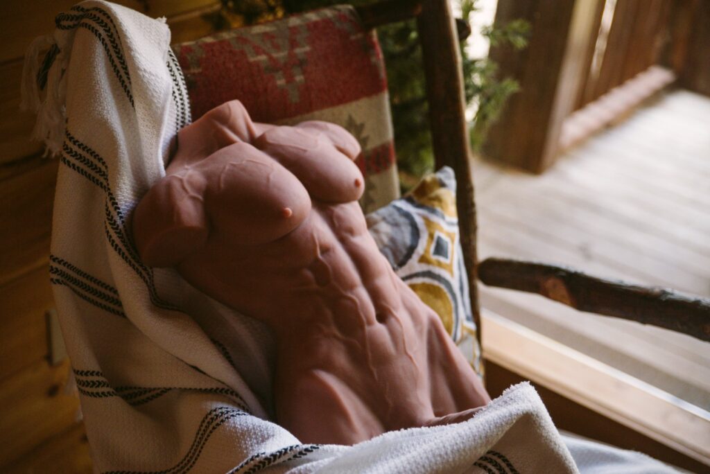 A male sex doll sits out on a cabin rocking chair, a camping blanket lewdly draped over the long, erect shaft, hiding it. For my How to Use a Male Sex Doll article.