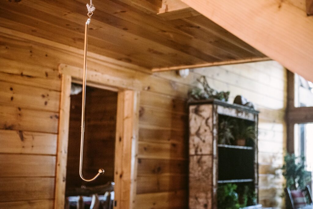 The Lockink anal hook hangs from the ceiling in a brown, wooden cabin. It stands out against the brown coloration behind it. It looks very shiny.