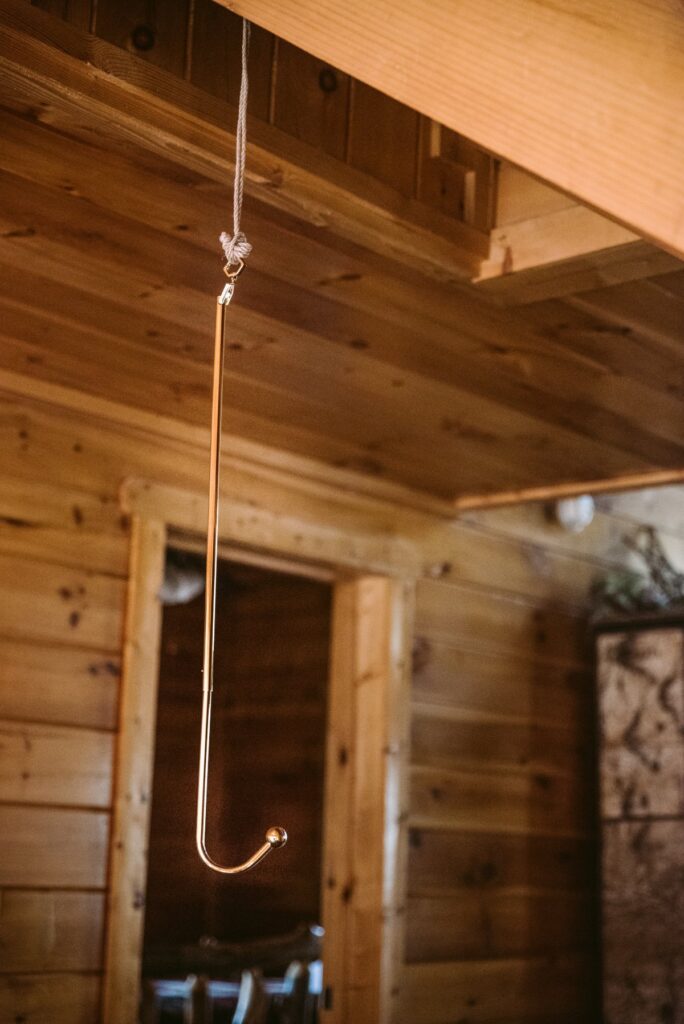 The Lockink anal hook hangs from the ceiling in a brown, wooden cabin. It stands out against the brown coloration behind it. It looks very shiny.