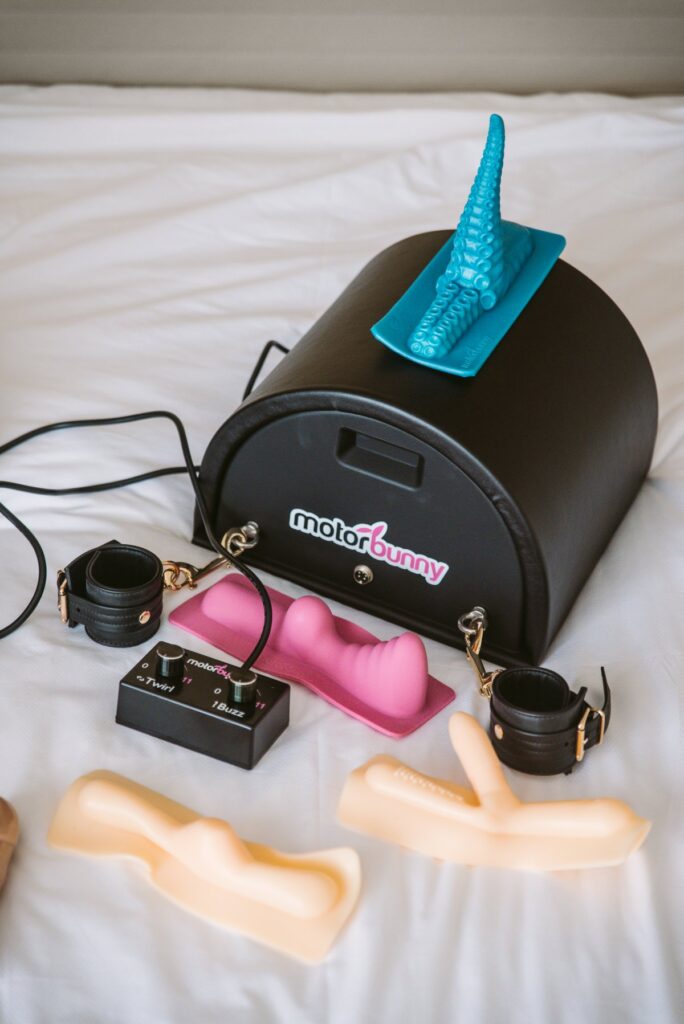 A Motorbunny sex machine sits out on a bed, surrounded by attachments, rope, bondage cuffs, and the remote control to control it. For my Kinky Ways to Use a Saddle Sex Machine article.