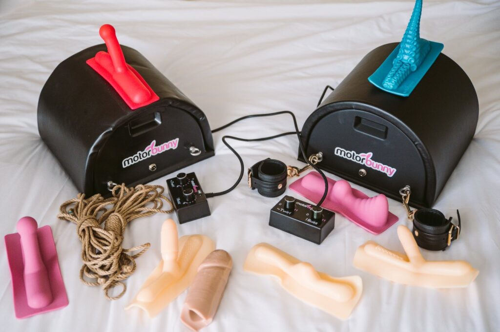 A Motorbunny sex machine sits out on a bed, surrounded by attachments, rope, bondage cuffs, and the remote control to control it. For my Kinky Ways to Use a Saddle Sex Machine article.