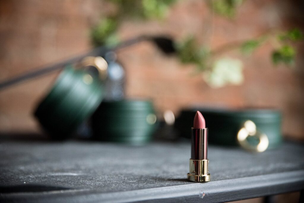 A tube of lipstick is sitting on a black table in the foreground. In the background, there are green bondage cuffs and a bondage collar. For my Hot AF Femdom Kissing Ideas article.