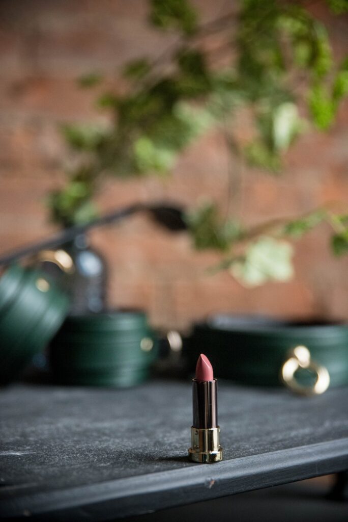 A tube of lipstick is sitting on a black table in the foreground. In the background, there are green bondage cuffs and a bondage collar. For my Hot AF Femdom Kissing Ideas article. 