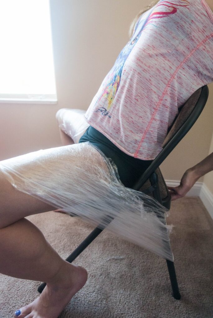A person is seated on a folding chair. Their legs are spread really far apart, and cling wrap is securing each thigh to each side of the chair, forcing the thighs to stay spread apart. For my plastic wrap bondage positions article. 