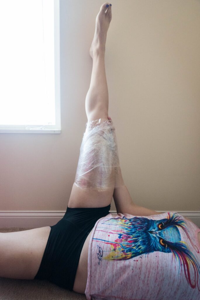 A person is laying on their back. One of their forearms is laying on top of their leg, which is pointed straight in the air against a wall. The forearm and the thigh are saran wrapped together. For my plastic wrap bondage positions article. 