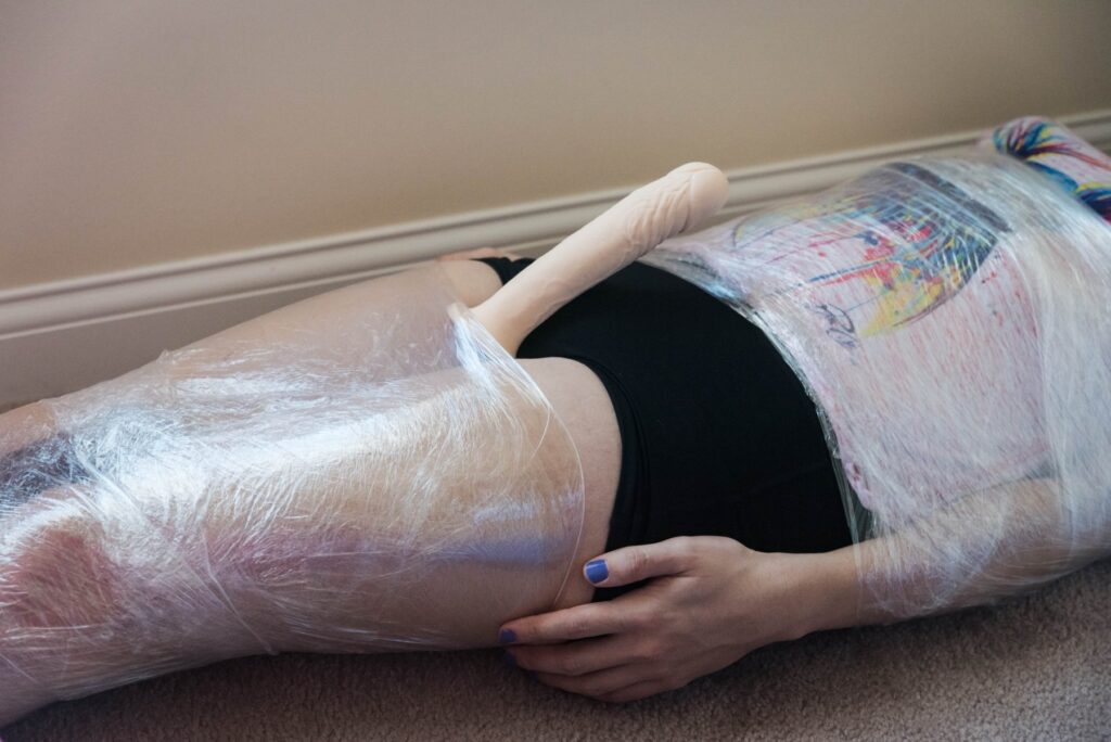A person is laying on their back. Their thighs are held together with cling wrap and their stomach and arms are forced close together with different layers of cling wrap. The hips are left entirely bondage-free. A dildo is sticking away from the person's hips, showcasing how easily accessible a penis would be here. For my plastic wrap bondage positions article. 