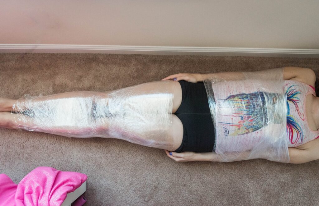 A person is laying on their back. Their thighs are held together with cling wrap and their stomach and arms are forced close together with different layers of cling wrap. The hips are left entirely bondage-free. For my plastic wrap bondage positions article. 
