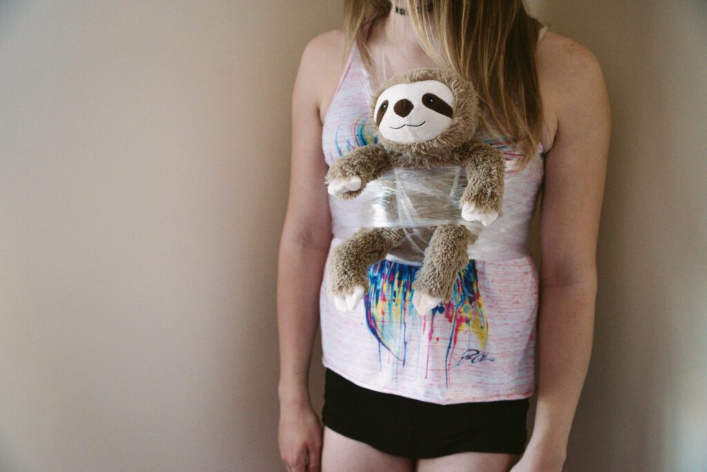 A person is standing. A small sloth stuffed animal is hanging from their chest from the cling wrap make-shift harness that we've wrapped around their chest and trapped the stuffed animal in. For my plastic wrap bondage positions article. 