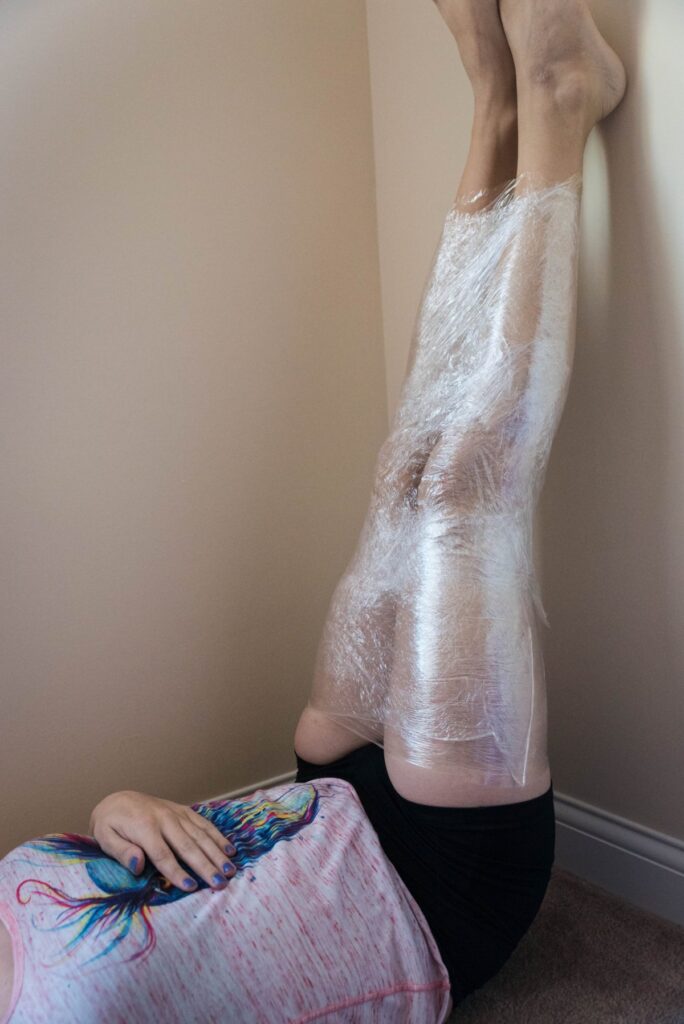 A person lays on their back. Their ankles are propped up against the wall, almost at a 90-degree angle. From hip to ankle, both legs are secured together with cling wrap. 
