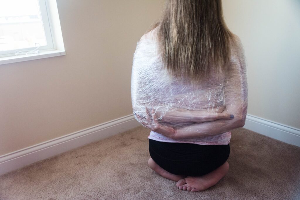 A person is kneeling, facing a wall. Their arms are behind their back, crossed over one another comfortably, and saran wrap has wrapped all around the entire upper body, keeping the arms positioned there. For my plastic wrap bondage positions article. 