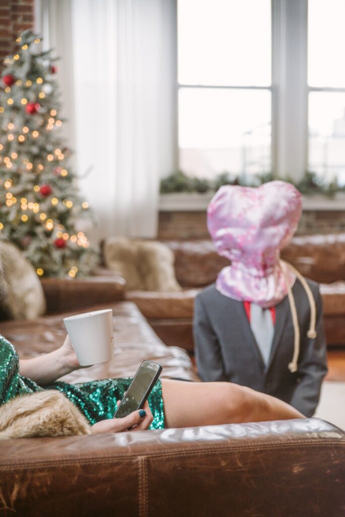 A person in a pink, black-out hood that doesn't allow them to see is kneeling in front of a couch. A different person is sitting in a nice dress, playing on their cell phone, comfortably on the couch. For my What is an Ignore Kink article. 