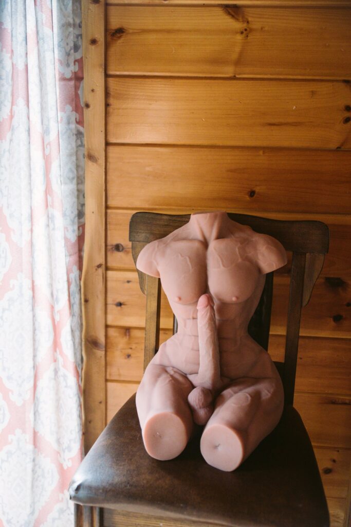 The naked sex doll sits on a chair, next to a window. The dildo penis on the front of the sex doll looks huge, even taller than the doll's nipples sitting in this position. For my ppunson Jax male sex doll review.