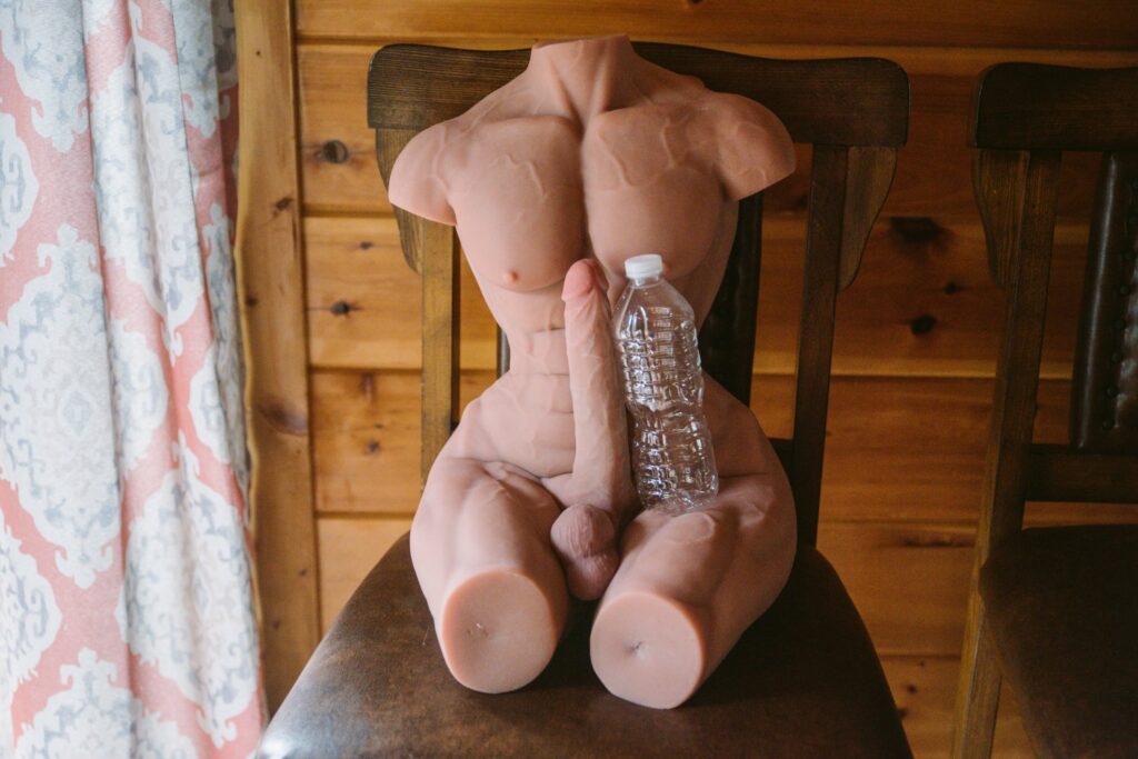 The naked sex doll sits on a chair, next to a window. The penis on the sex doll is shown next to an empty water bottle. They look to be about the same height, showcasing how the dildo just looks gigantic because it's attached to a smaller-than-average torso. For my ppunson Jax male sex doll review.