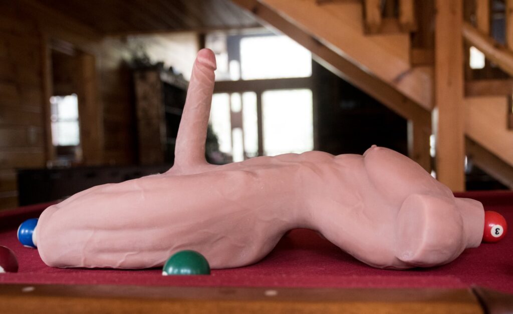 The sex doll lays, back-down, on a pool table. This photo is taken from the side, showcasing how tall the dildo protrudes away from the sex doll itself. The dildo stands up by itself without any help. For my ppunson Jax male sex doll review.