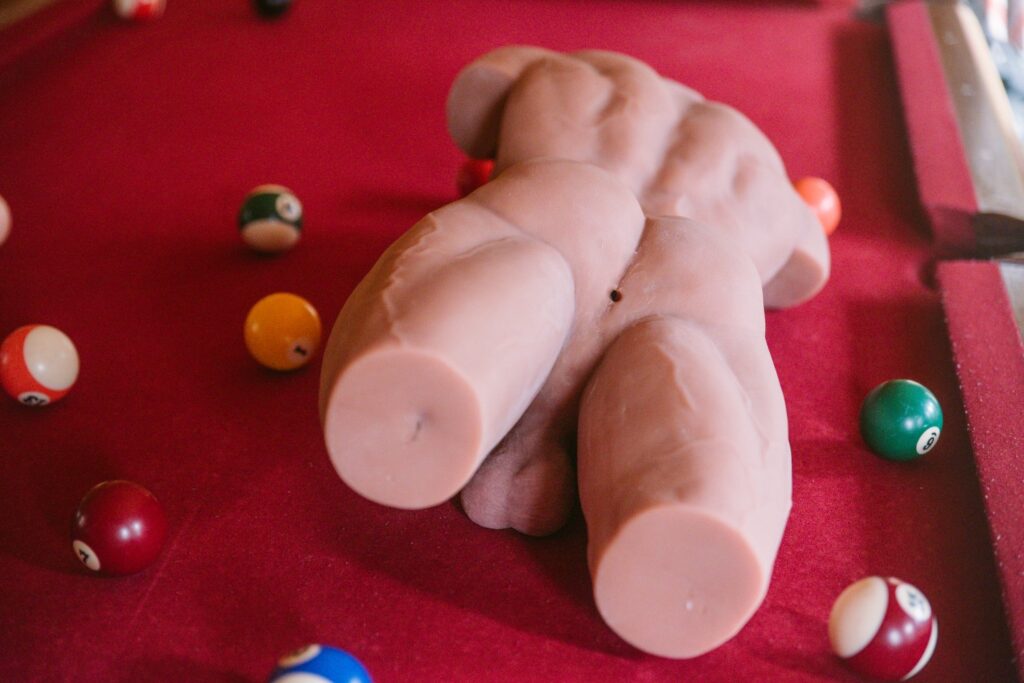 The sex doll lays, stomach-down, on top of a pool table. The huge, hard dildo is resting underneath of it, providing a "see-saw" of the weight dispersion of the sex doll. The doll is always off-center on the right or left when left "at rest".