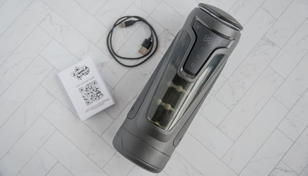 The Fleshy Thrust Sync lays flat on a tiled background, shown next to the charging cable and the instruction booklet it comes with. For my Fleshy Thrust Sync Review.