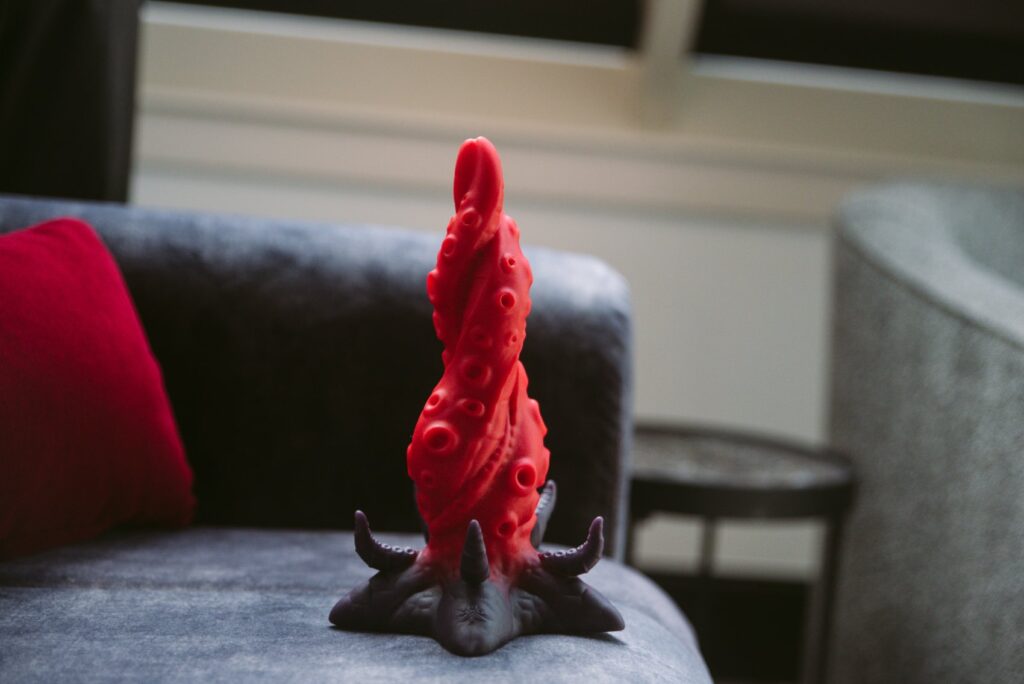 The Starfish dildo sits on a couch. The room is dark behind it, really highlighting the colors and unique shape of the dildo. For my Juliet Toys Starfish dildo review.