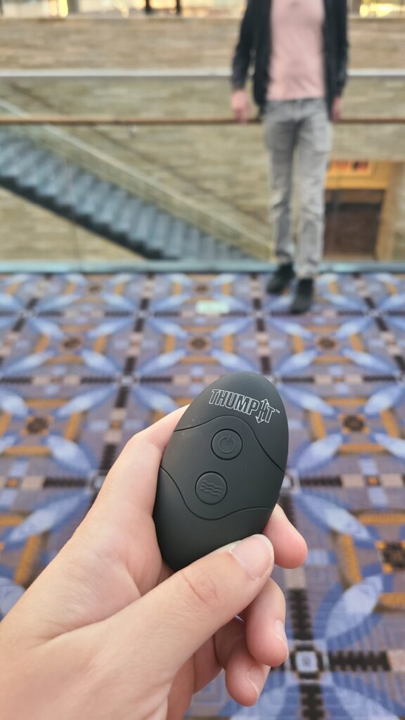 Close-up on a sex toy remote control which is held in my hand. It's pointing at the crotch of my submissive who is standing in the background of the shot in the middle of a conference center. Article for my scenes for public femdom teasing article. 