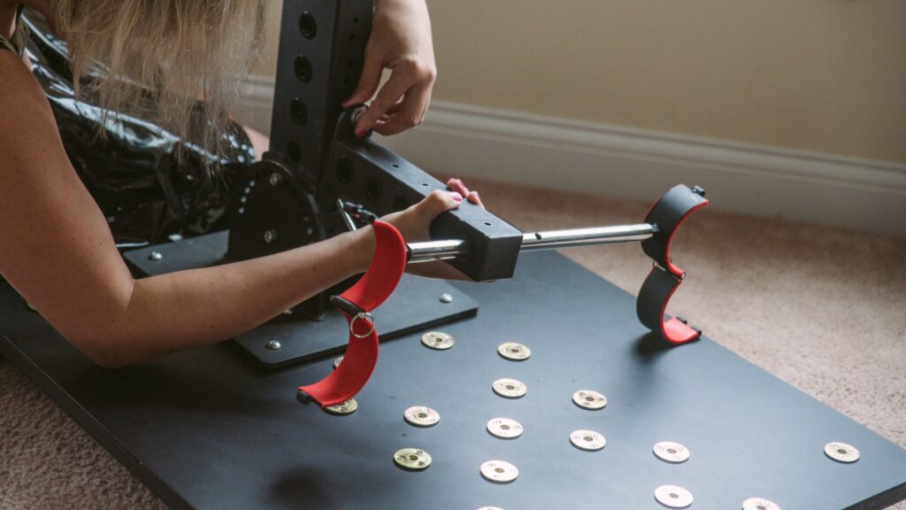 My partner putting the ankle cuff post onto the L-shaped frame. One of her hands has to support the weight of the post while the other tries to fit a bolt through two different holes, showcasing how all of the modular pieces fit together. For my Fetdom Multi-Position Floating Stockade Pillory Review.