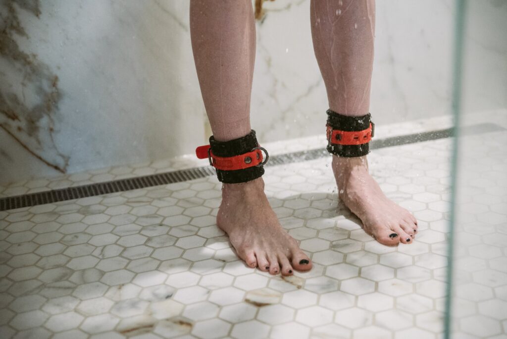A person stands in the shower, wearing silicone black and red bondage cuffs. Water is dripping down their legs. For my Bondage omorashi tips to minimize mess.