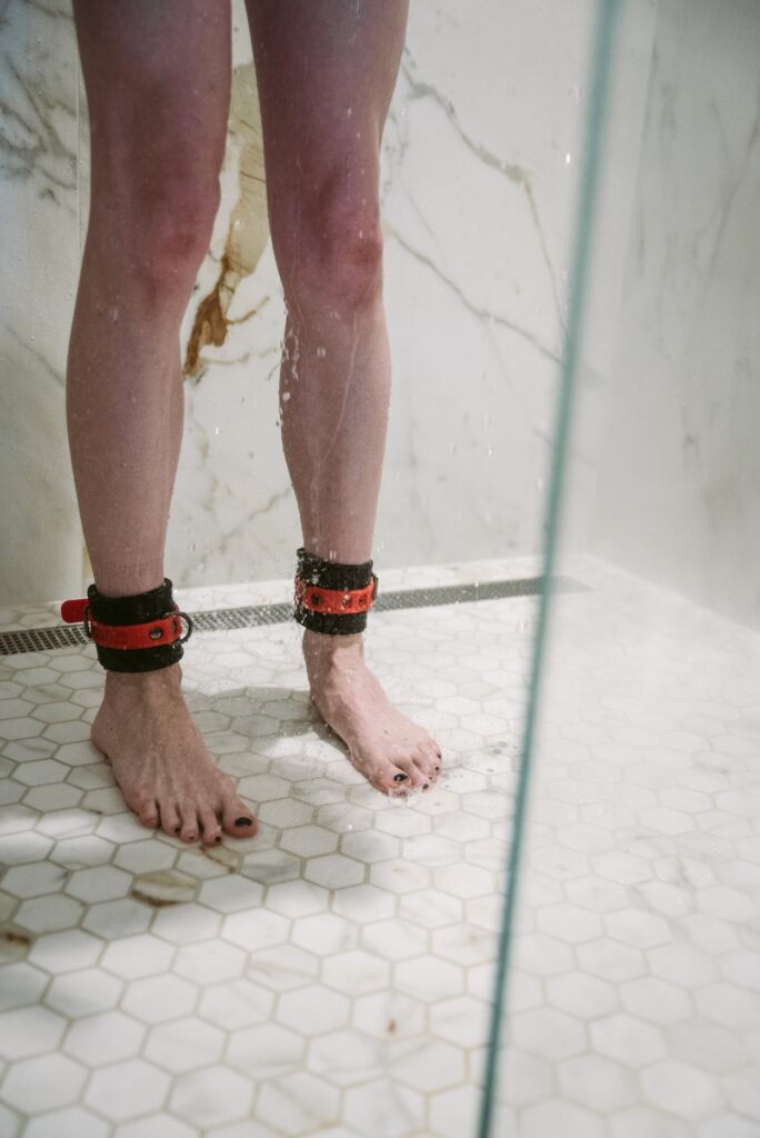 A person stands in the shower, wearing silicone black and red bondage cuffs. Water is dripping down their legs. For my Bondage omorashi tips to minimize mess.