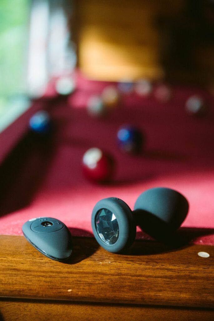 The b-Vibe plug sits on the edge of a pool table with pool table balls spread out on the table. The remote to control the plug sits besides it. For my b-Vibe XXL Vibrating Jewel Plug Review.