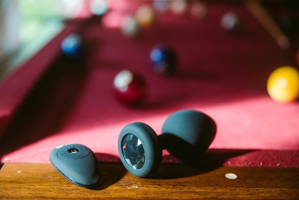 The b-Vibe plug sits on the edge of a pool table with pool table balls spread out on the table. The remote to control the plug sits besides it. For my b-Vibe XXL Vibrating Jewel Plug Review.