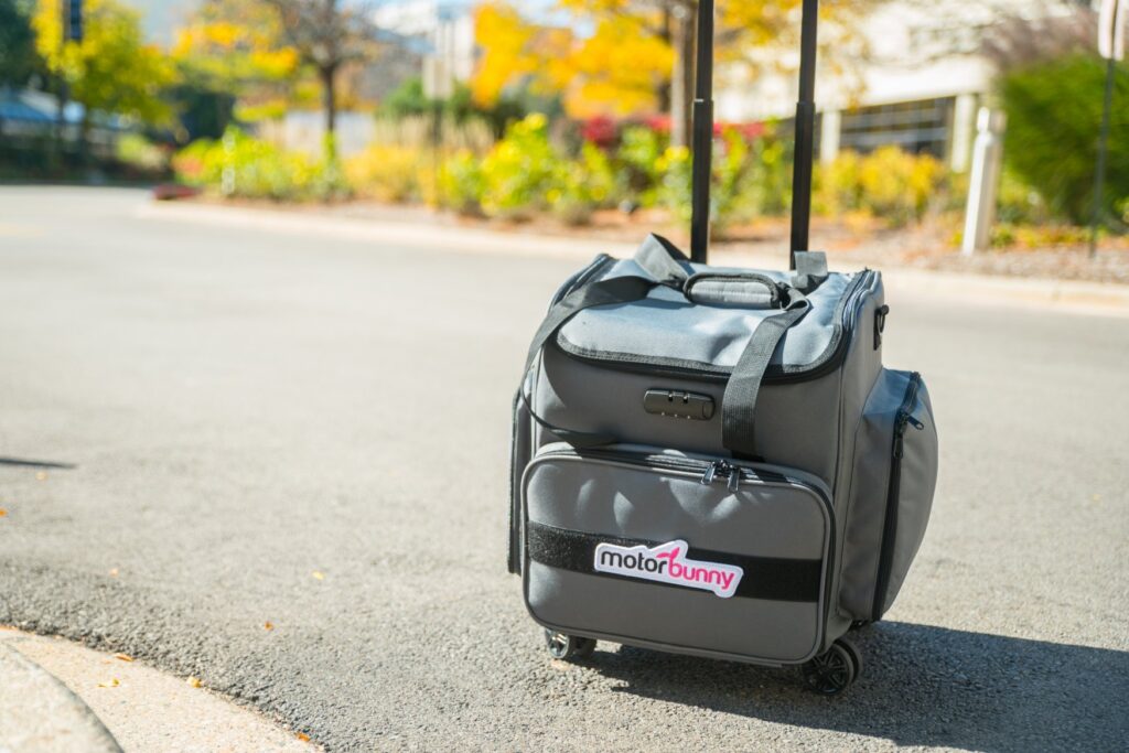 The Motorbunny rolling luggage case is sitting outside on the sidewalk. There's beautiful fall trees in the background. For my how to travel with BDSM toys article.