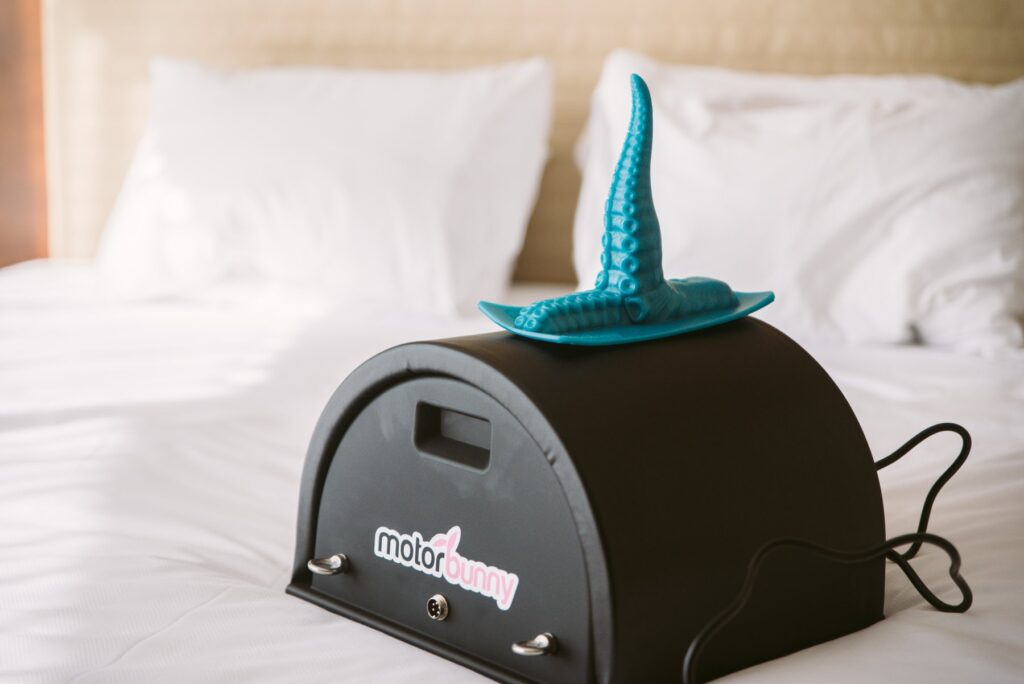 A Motorbunny sex machine sits out on a bed. The bed looks soft and welcoming. A tentacle dildo is strapped to the top of the Motorbunny machine. For my Sybian for Male? article.